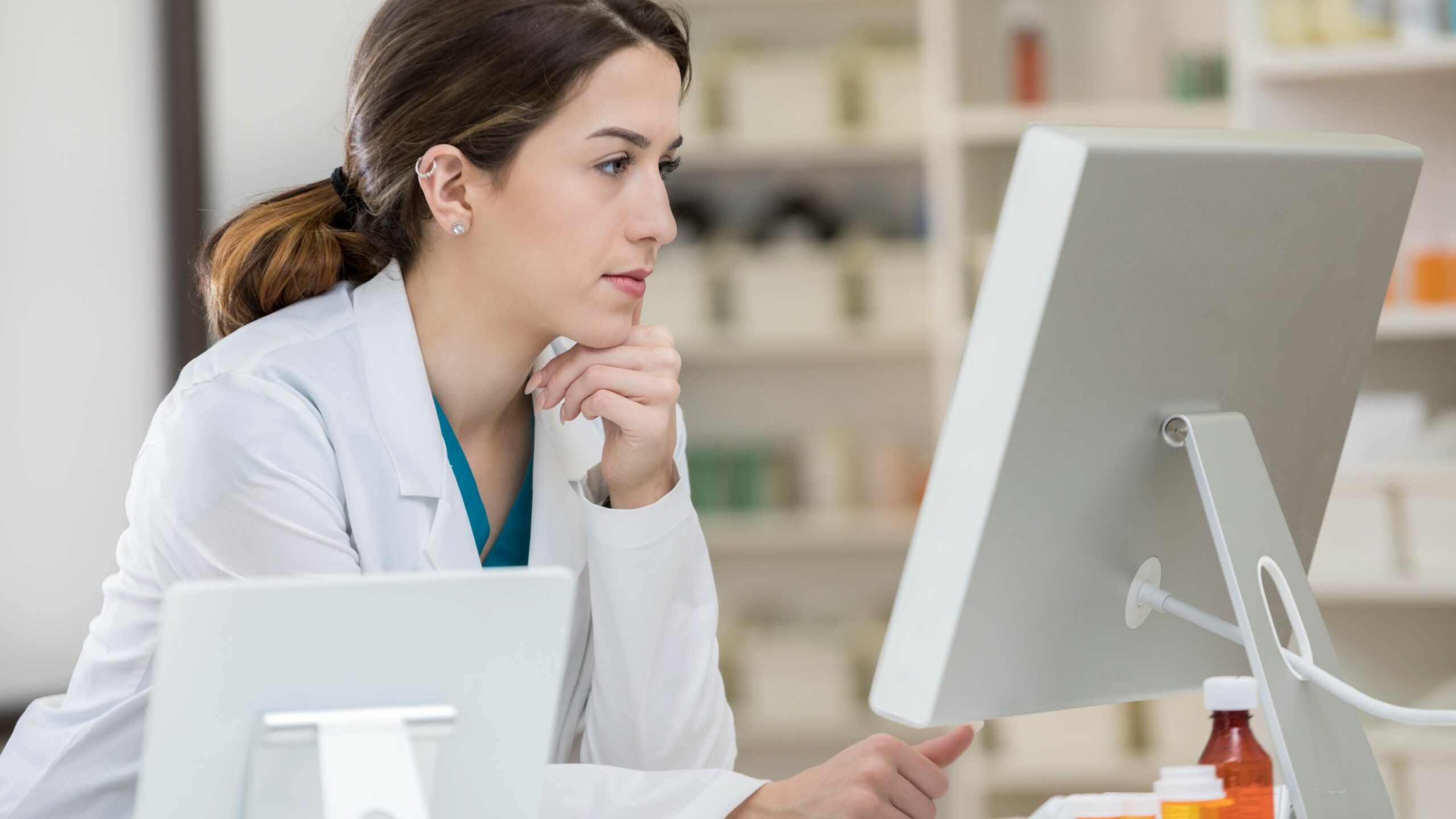 A serious Caucasian pharmacist leans on her hand at a table as she studies the latest incoming prescriptions on a computer screen.  There is pharmaceutical equipment in the foreground and shelves in the background.
