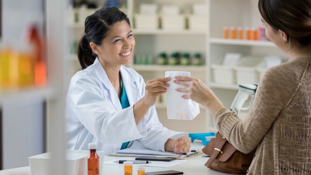 Friendly mid adult Asian female pharmacist hands a paper bag to young female customer at the checkout counter.